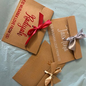 May include: Three brown paper envelopes with gold, silver, and red ribbons tied around them. The envelopes have the text "EID MUBARAK LOVE FROM US!" printed on them, along with the names "Jasmina", "Radiyah", and "Jasmina".