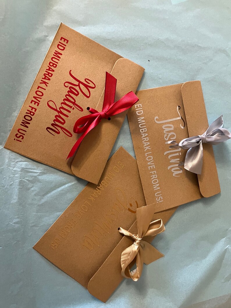 May include: Three brown paper envelopes with gold, silver, and red ribbons tied around them. The envelopes have the text "EID MUBARAK LOVE FROM US!" printed on them, along with the names "Jasmina", "Radiyah", and "Jasmina".