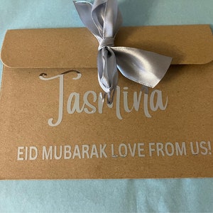 May include: A brown paper envelope with a silver ribbon tied in a bow. The envelope has the name "Jasmina" written on it in silver lettering. The text "Eid Mubarak Love From Us!" is also written on the envelope in silver lettering.