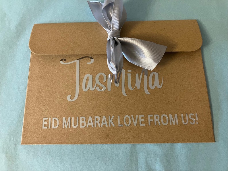 May include: A brown paper envelope with a silver ribbon tied in a bow. The envelope has the name "Jasmina" written on it in silver lettering. The text "Eid Mubarak Love From Us!" is also written on the envelope in silver lettering.