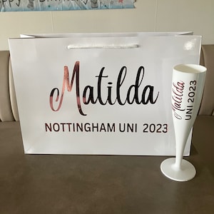 May include: A white gift bag and champagne flute with rose gold text that reads "Matilda" and "Nottingham UNI 2023". The flute also has "Matilda UNI 2023" printed on it. A banner that says "Happy Birthday" is in the background.