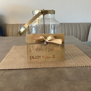 May include: A brown envelope with a gold ribbon and the text "Eid Mubarak" and "Enjoy Your Gift" in gold lettering.