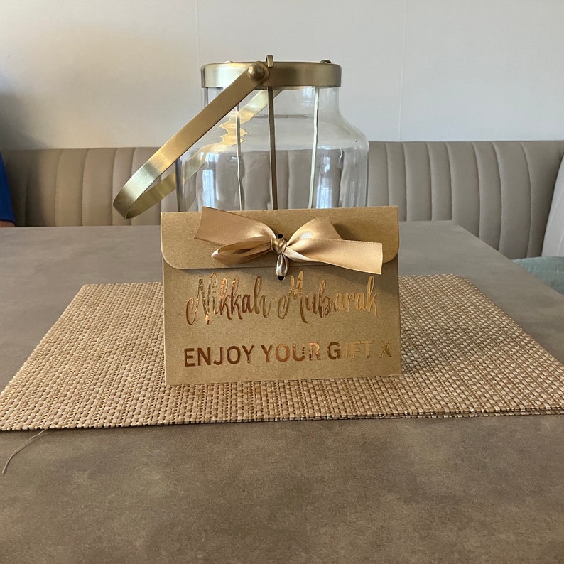 May include: A brown envelope with a gold ribbon and the text "Eid Mubarak" and "Enjoy Your Gift" in gold lettering.
