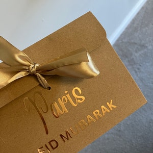 May include: A brown paper envelope with a gold ribbon tied around it. The envelope has gold lettering that says "Paris" and "Eid Mubarak".