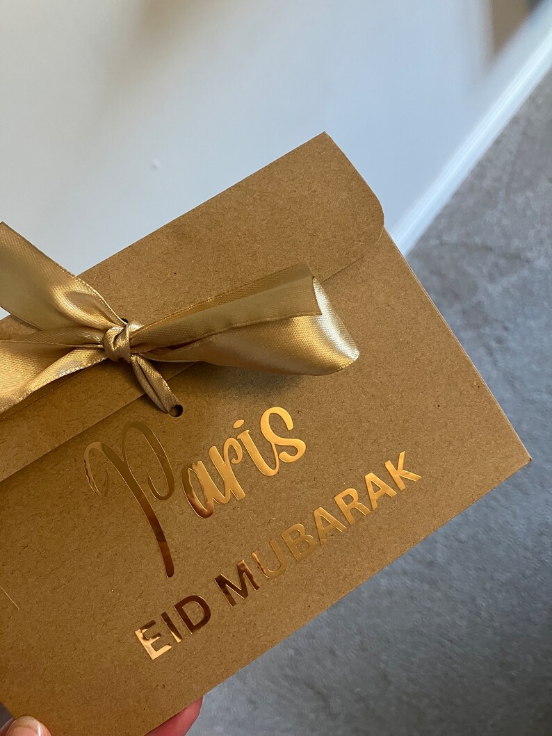 May include: A brown paper envelope with a gold ribbon tied around it. The envelope has gold lettering that says "Paris" and "Eid Mubarak".
