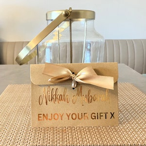 May include: A brown paper envelope with a gold ribbon tied in a bow. The envelope has the text "Nikkah Mubarak" and "Enjoy Your Gift X" printed in gold foil.