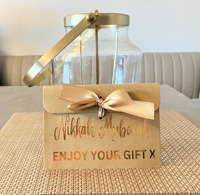 May include: A brown paper envelope with a gold ribbon tied in a bow. The envelope has the text "Nikkah Mubarak" and "Enjoy Your Gift X" printed in gold foil.