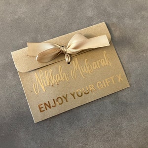 May include: A brown paper envelope with a gold ribbon tied in a bow. The envelope has the text "Nikkah Mubarak" and "Enjoy Your Gift X" printed in gold.