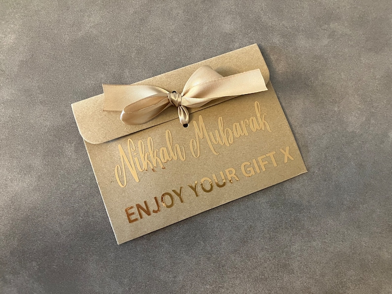 May include: A brown paper envelope with a gold ribbon tied in a bow. The envelope has the text "Nikkah Mubarak" and "Enjoy Your Gift X" printed in gold.