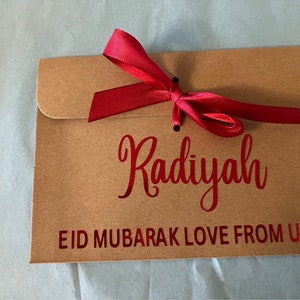 May include: A brown paper envelope with a red ribbon tied around it. The envelope has the name "Radiyah" written on it in red lettering. The envelope also has the text "Eid Mubarak Love From Us!" written on it in red lettering.