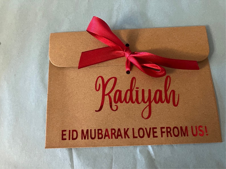 May include: A brown paper envelope with a red ribbon tied around it. The envelope has the name "Radiyah" written on it in red lettering. The envelope also has the text "Eid Mubarak Love From Us!" written on it in red lettering.