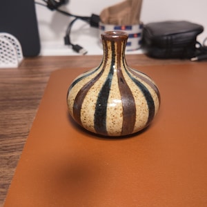 Op de afbeelding: Een kleine, bolvormige keramische vaas met een smalle hals en een uitlopende rand. Het lichaam heeft een gespikkelde beige basis met afwisselende verticale strepen in donkerbruin en zwart, wat een opvallend patroon vormt van de voet tot de hals.