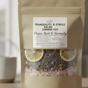 May include: A kraft paper bag labeled "Tranquility & Stress Relief Simmer Pot" with the words "Peace, Rest & Serenity." The bag contains lavender, chamomile, pink salt, and two lemon slices. A white mug is visible in the background.