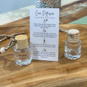 May include: Two small, clear glass car air freshener bottles with wooden lids and attached strings sit on a wooden tray. One bottle is labeled "Warm Vanilla." A card provides instructions for use. A decorative container is in the background.