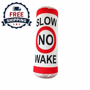 May include: A cylindrical white cushion featuring a red circular border and the black text SLOW NO WAKE printed vertically. The design mimics a nautical speed limit sign with red horizontal bands at the top and bottom of the fabric surface.