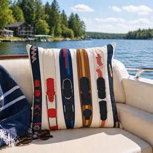 May include: A cushion with a cream background featuring a pattern of vintage water skis in various colours. The skis include red, blue, and brown designs. The cushion is on a boat with a lake and trees in the background.