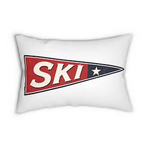 Op de afbeelding: Een rechthoekig wit sierkussen met een gecentreerde driehoekige wimpelafbeelding. De wimpel is rood met een donkerblauwe punt en toont de witte tekst "SKI" in een vetgedrukt vintage lettertype, vergezeld van een kleine witte ster op het blauwe deel.