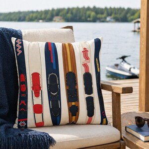 May include: Cushion with a cream background featuring a pattern of water skis in various colours, including red, blue, and brown. The cushion is on a wooden chair with a lake and trees in the background.