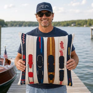 May include: A cushion with a design of vintage water skis in red, blue, and brown. The cushion has a cream-coloured background and is held by a person wearing a navy blue hat with the text "LAKE LIFE".