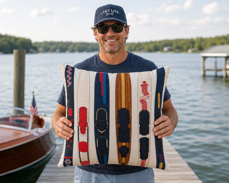 May include: A cushion with a design of vintage water skis in red, blue, and brown. The cushion has a cream-coloured background and is held by a person wearing a navy blue hat with the text "LAKE LIFE".
