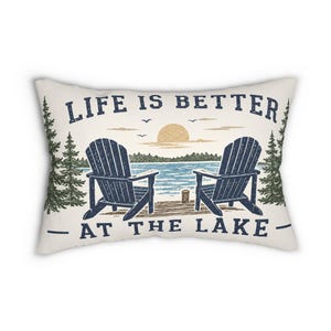May include: A rectangular off-white throw pillow featuring a graphic of two dark blue Adirondack chairs on a wooden dock overlooking a lake. The text reads LIFE IS BETTER AT THE LAKE in dark blue letters, with green pine trees and a sun.