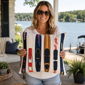 May include: Cream rectangular cushion featuring a pattern of vintage water skis in red, navy blue, and brown. The skis are arranged vertically, creating a decorative design. The cushion is held by a person.