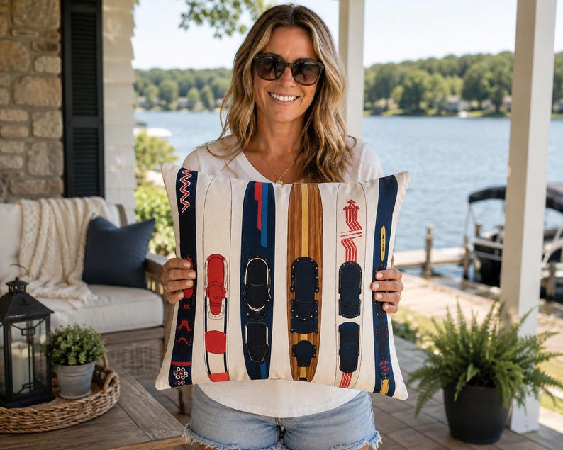 May include: Cream rectangular cushion featuring a pattern of vintage water skis in red, navy blue, and brown. The skis are arranged vertically, creating a decorative design. The cushion is held by a person.
