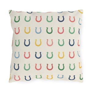 May include: Square throw pillow with a repeating pattern of colorful horseshoes in shades of green, blue, yellow, and pink on a cream background. The pillow is a decorative item, suitable for a living room or bedroom.