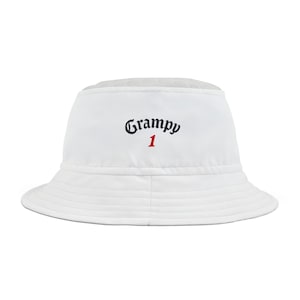 May include: White bucket hat featuring the word "Grampy" in black script and the number "1" in red. The hat has a wide brim and is made of a smooth material. A casual hat for sun protection, suitable for various outdoor activities.