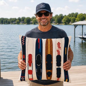 May include: A rectangular cushion with a cream-coloured background, decorated with vertical images of vintage water skis in shades of red, blue, and brown. The cushion is likely made of a soft fabric and would suit a nautical-themed room.