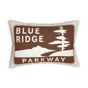 Può includere: Un cuscino rettangolare con sfondo color crema e un design grafico marrone. Il motivo presenta la scritta bianca "BLUE RIDGE PARKWAY" accanto alla sagoma bianca di una catena montuosa, una strada tortuosa e un alto albero di pino.