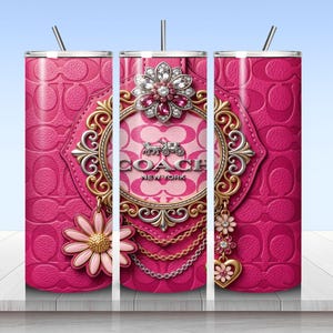 May include: A cylindrical tumbler with a pink leather-textured background featuring a repeating logo pattern. The design includes a central ornate gold and silver frame with the text COACH NEW YORK, a large jeweled flower, and hanging charms.