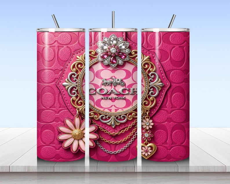 May include: A cylindrical tumbler with a pink leather-textured background featuring a repeating logo pattern. The design includes a central ornate gold and silver frame with the text COACH NEW YORK, a large jeweled flower, and hanging charms.