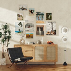 Pode incluir: Um interior de sala de estar com um armário de madeira, uma poltrona preta e um abajur de pé com duas cúpulas circulares brancas. A parede é decorada com uma coleção de obras de arte emolduradas que representam paisagens marinhas, paisagens e figuras.