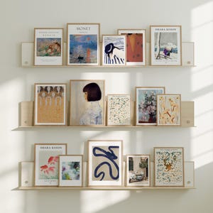 May include: A collection of framed art prints displayed on three wooden shelves. The prints feature various styles, including floral, abstract, and landscape paintings. Some prints have text, including "MONET" and "OHARA KOSON". The frames are a light wood color.