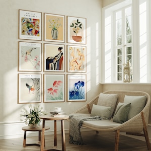 Op de afbeelding: Een woonkamer met een witte bank en een muur die is versierd met ingelijste kunstprints. De prints tonen een verscheidenheid aan onderwerpen, waaronder vogels, bloemen en abstracte ontwerpen. De prints zijn in een rasterpatroon op de muur gerangschikt. De bank is gemaakt van riet en heeft een wit kussen. Er staat een kleine tafel voor de bank met een kopje en schotel erop.