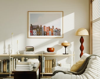 May include: A living room interior with a white couch, a wooden bookshelf, and a framed print of a row of colorful houses. The print is hanging on a white wall. A wooden lamp with a white shade is on the floor next to the couch.