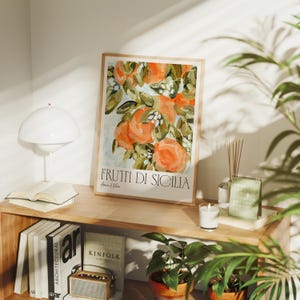 Könnte beinhalten: Ein gerahmter Druck mit einem Gemälde von orangefarbenen Zitrusfrüchten an einem Zweig mit grünen Blättern. Der Text "FRUTTI DI SICILIA" ist in Schwarz am unteren Rand des Drucks gedruckt.