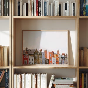May include: A framed print featuring a row of colorful buildings with chimneys. The buildings are painted in shades of red, orange, yellow, blue, and white. The print is displayed on a wooden shelf surrounded by books.