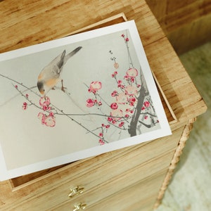 May include: A delicate watercolor painting of a small bird perched on a branch of a flowering tree. The bird is light brown with a white breast and is depicted pecking at a pink blossom. The tree is in full bloom with pink flowers and green leaves. The painting is framed and displayed on a wooden dresser.
