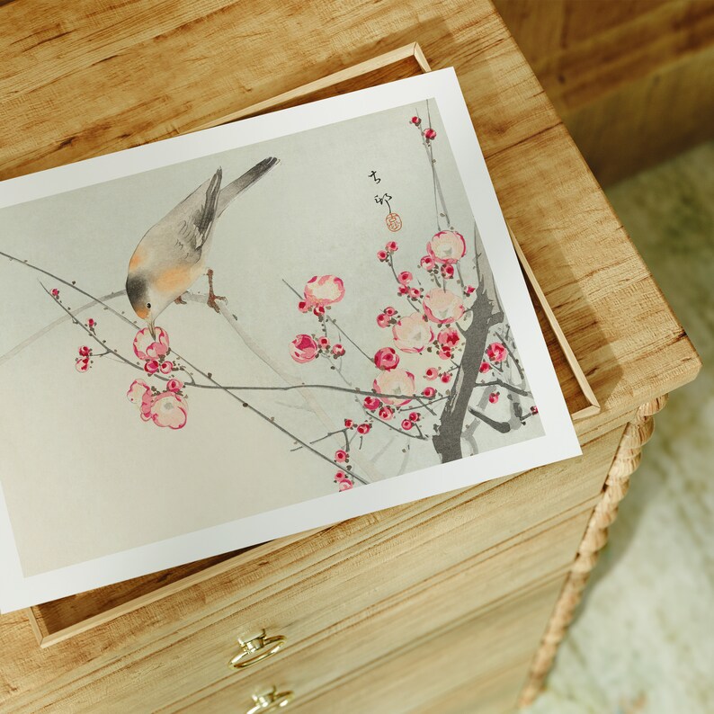 May include: A delicate watercolor painting of a small bird perched on a branch of a flowering tree. The bird is light brown with a white breast and is depicted pecking at a pink blossom. The tree is in full bloom with pink flowers and green leaves. The painting is framed and displayed on a wooden dresser.