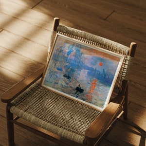 May include: A framed art print of an Impressionist painting, featuring a sunrise over a harbor. The artwork displays a vibrant palette of blues, oranges, and reds, with small boats and buildings in the background. The print is displayed on a wooden chair.