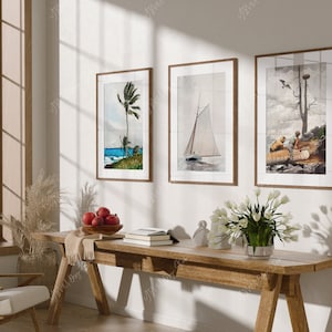 Peut inclure: Trois impressions d'art encadrées accrochées à un mur blanc au-dessus d'une table en bois. Les impressions représentent un voilier, un palmier et une scène avec deux personnes dans un bateau. La table est en bois et a une finition rustique. Il y a un bol de pommes rouges sur la table.