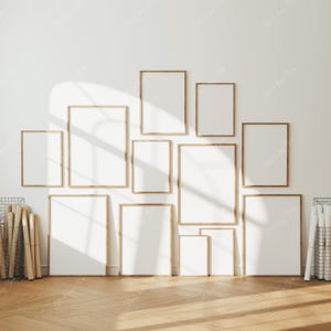 May include: A collection of framed art prints in various sizes, arranged on a white wall. The frames are a light wood color, and the prints are blank, ready for artwork or photos. The room has a wooden floor and is well-lit.