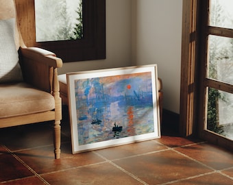 May include: A framed print of a painting depicting a seascape with boats and a red sun setting over the water. The painting is in a light wood frame and is hanging on a wall.