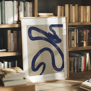 Puede incluir: Impresión artística enmarcada con un diseño abstracto. Presenta un dibujo lineal azul marino sobre un fondo beige, enmarcado en madera. La obra se apoya en una estantería llena de libros, creando una estética moderna y minimalista.