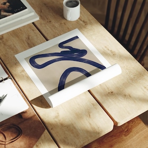 May include: An abstract art print featuring a beige background with a dark blue, flowing line design. The print is partially rolled, revealing the white backing. The artwork rests on a light-colored wooden surface, alongside a white mug and other items.