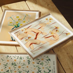 May include: Three framed prints with floral designs. The top print features a bouquet of white flowers in a blue and white vase on a light peach background. The middle print features a stylized abstract design of yellow flowers and peach-colored shapes on a cream background. The bottom print features a white floral pattern on a green background.
