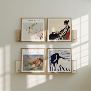 May include: Four framed art prints displayed on a light wood shelf. The prints feature various artistic styles, including a bird on a flowering branch, a figure in striped clothing, a Renoir painting, and a figure with a hat.