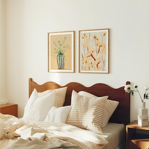 May include: Two framed art prints hanging on a white wall above a wooden headboard. The print on the left features a bouquet of white flowers in a blue and white striped vase. The print on the right features a group of figures in a field of yellow flowers.